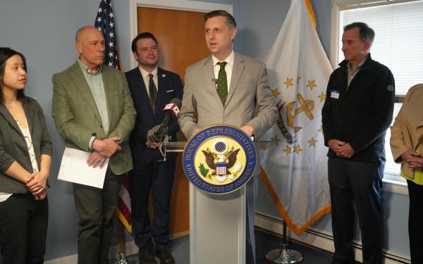 Magaziner speaks at a podium surrounded by leaders at South Kingstown Housing Authority