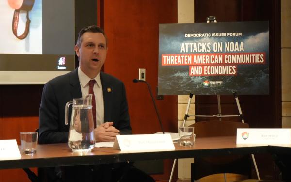 Magaziner is sitting, speaking next to a sign that reads 'Attacks on NOAA Threaten American Communities and Economies'